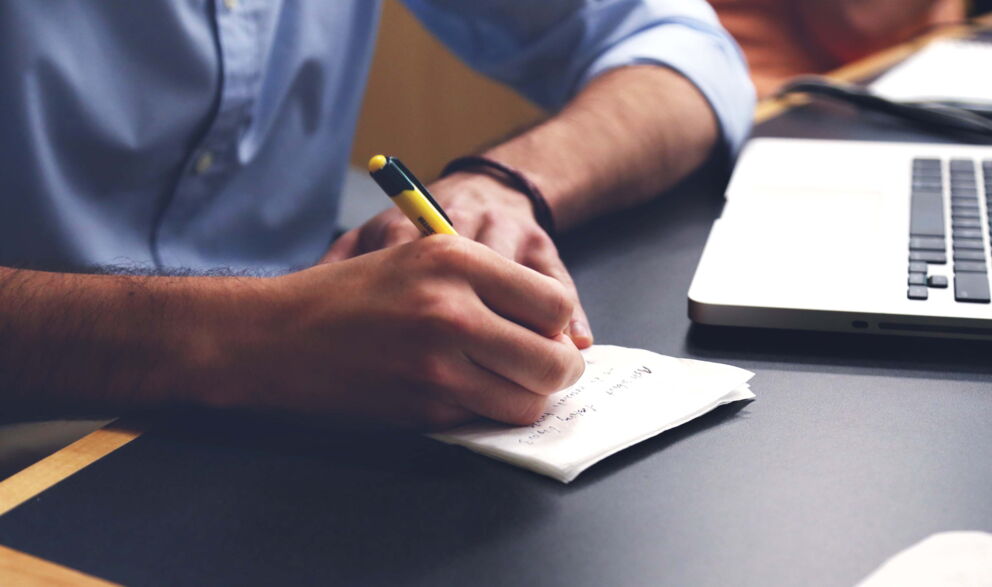 A man writes notes on a piece of paper next to his laptop